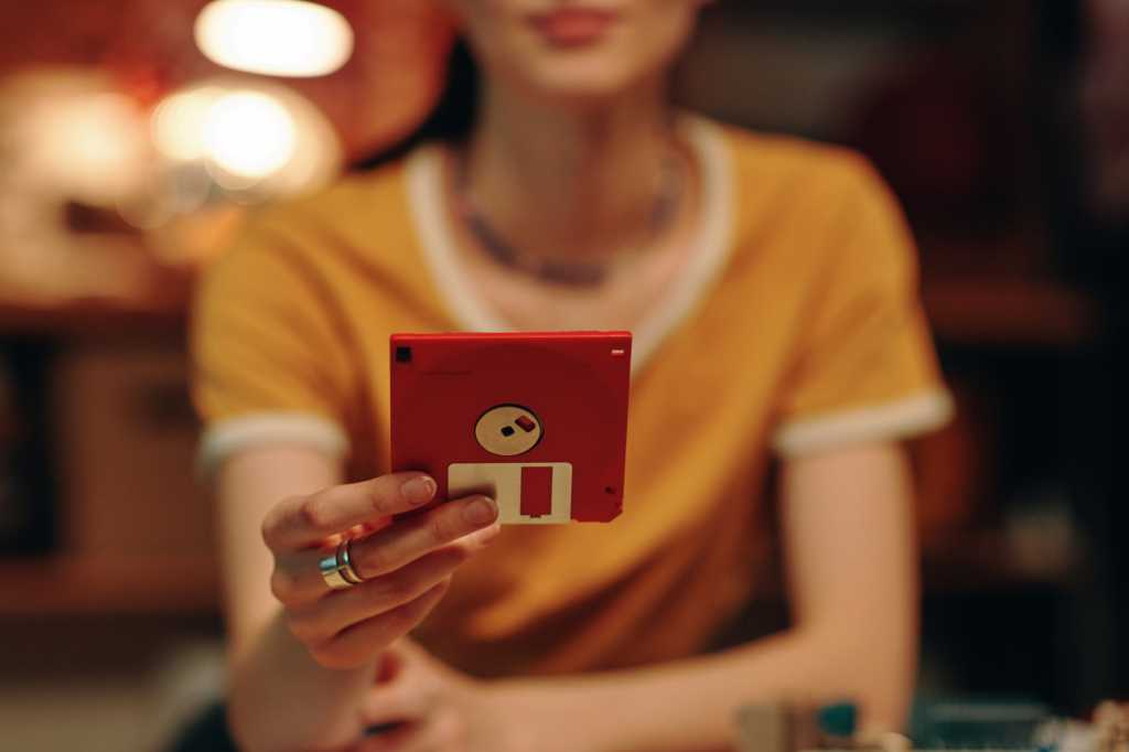 Woman In Yellow Holding Up A Red Floppy Disk At Her Desk.jpg