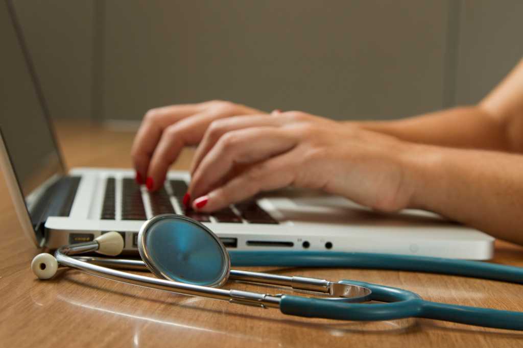 Woman On Laptop Keyboard With Stethoscope Nearby On Desk 1.jpg